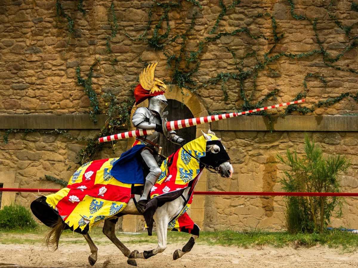 A knight in red and gold heraldic livery on horseback, galloping past a stone castle wall with a lance raised