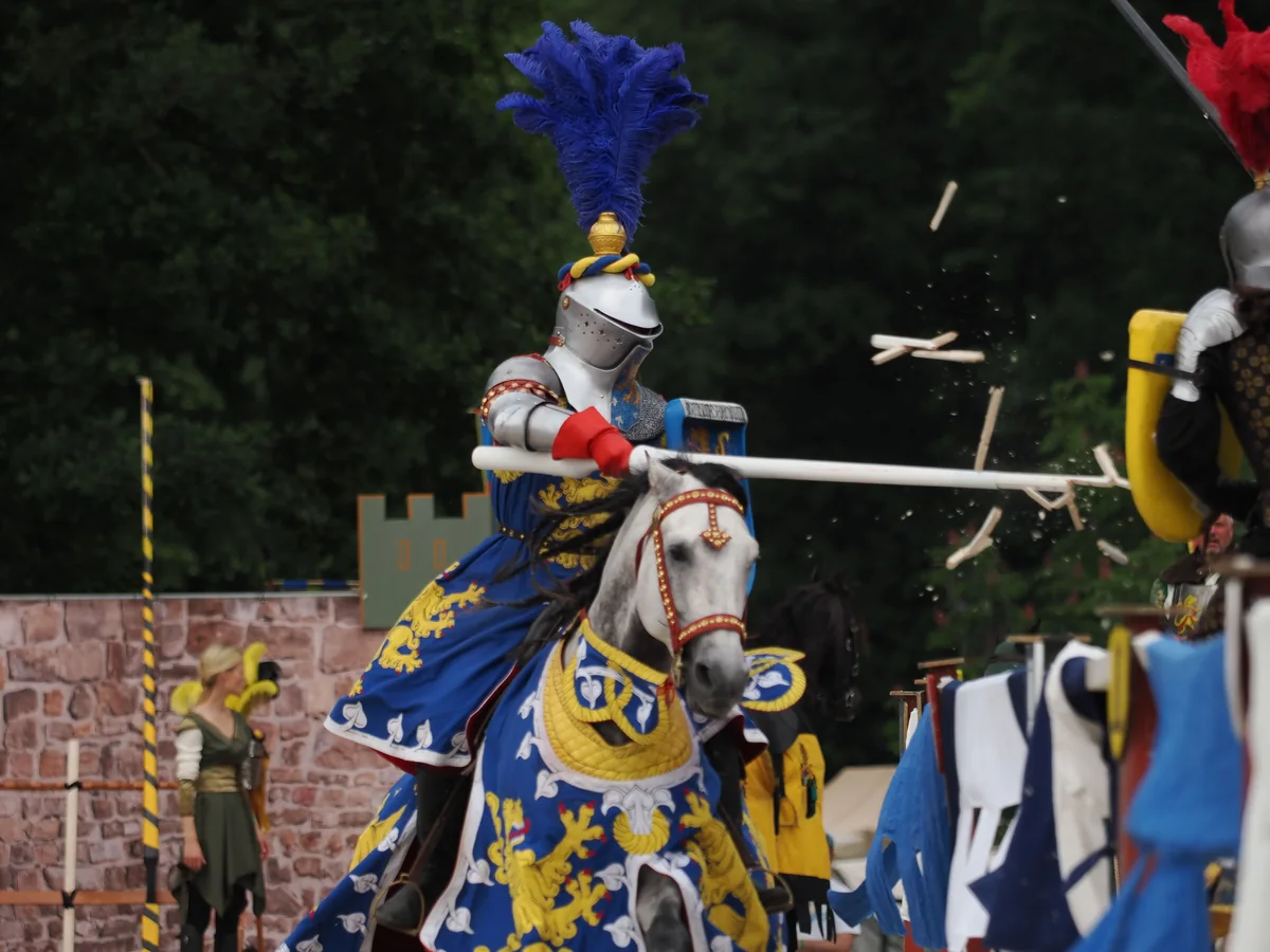 A mounted knight in blue and gold livery jousting, lance shattering on impact