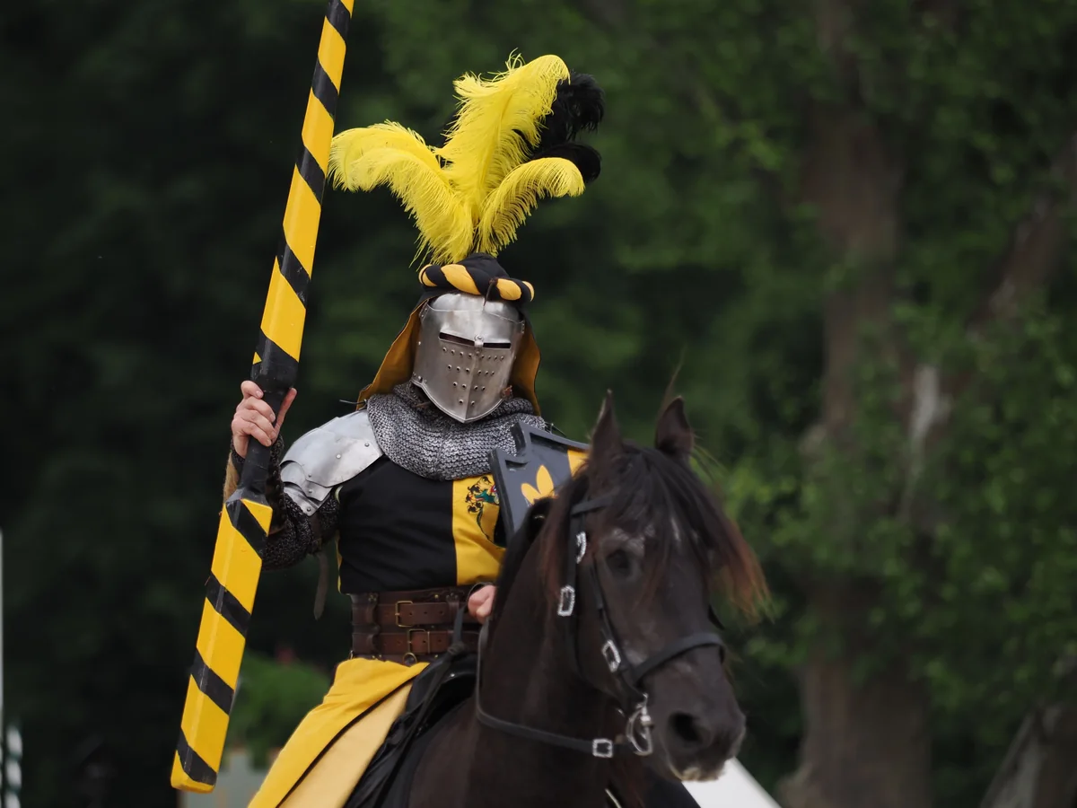 A mounted knight in black and yellow team livery holding a striped tournament lance upright